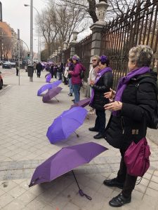 Women standing in a row in front of a building with purple umbrellas in front of them.